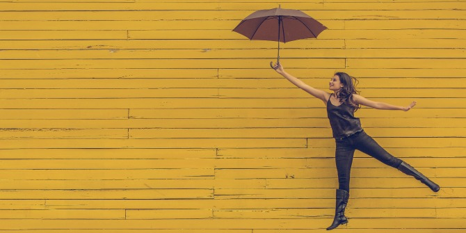 Woman against a yellow background with one leg up behind her holding an umbrella in her arm.